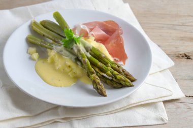 Yeşil kuşkonmaz, patates püresi, jambon ve hafif hollandez sosuyla servis edilir. Beyaz bir tabakta, ahşap bir masada. Kopya alanı, seçilmiş odak, dar bir tarla derinliği.