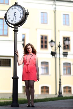 Pembe elbiseli çekici esmer, şehrin büyük saatine yerleştirildi. Güzel kız güneşli bir günde yürür. Saçları kısa pembe elbiseli genç bir kadın..