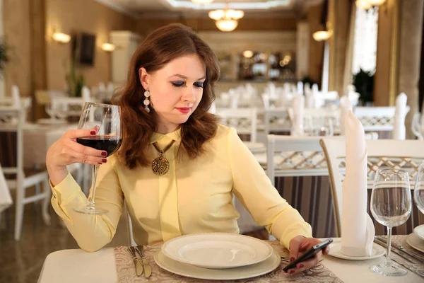 A young lady is looking at the phone in a cafe. Smokey eyes makeup and red ladies business manicure.Beautiful brunette is sitting in a restaurant with a glass of wine. The girl in the cafe.