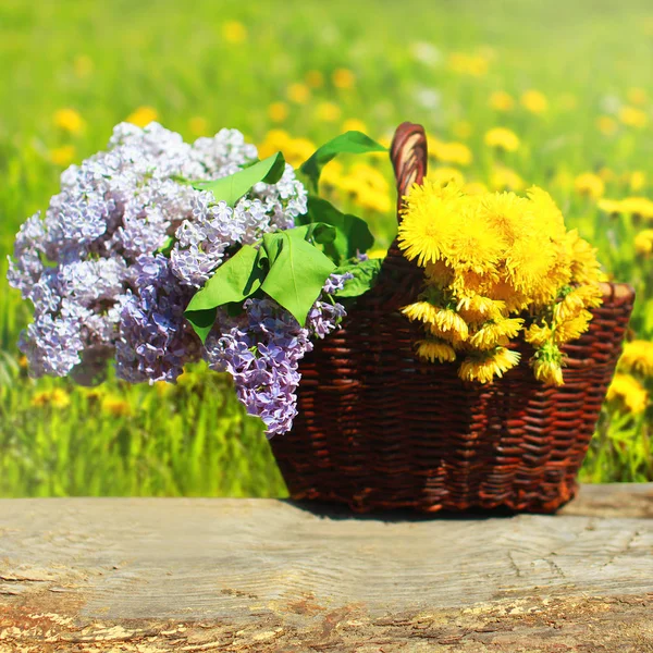 Summer wicker basket with yellow dandelions flowers and lilacs o — Stock Photo, Image