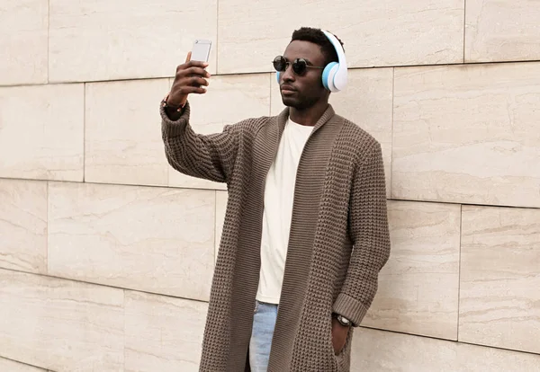 スタイリッシュなアフリカ人男性写真を撮るスマートフォンでワイヤレスヘッドフォンで音楽を聴く茶色のニットカーディガン 壁の背景の上の街のサングラスを身に着けている — ストック写真