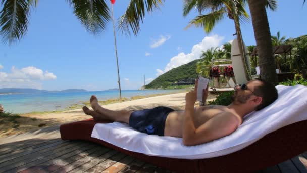 Человек читает книгу под кокосовой пальмой на тропическом пляже — стоковое видео