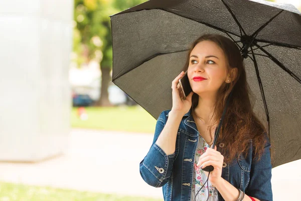 Mujer joven sosteniendo el paraguas y hablando en el teléfono celular al aire libre en el día lluvioso — Foto de Stock