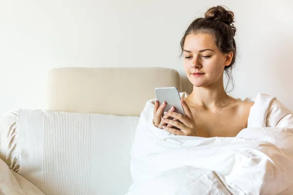 Mooie jonge vrouw vallende deken zit op bed met slimme telefoon — Stockfoto