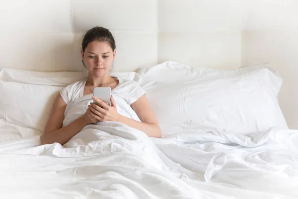 De mooie jonge meisje portret met slimme telefoon op bed in de ochtend — Stockfoto