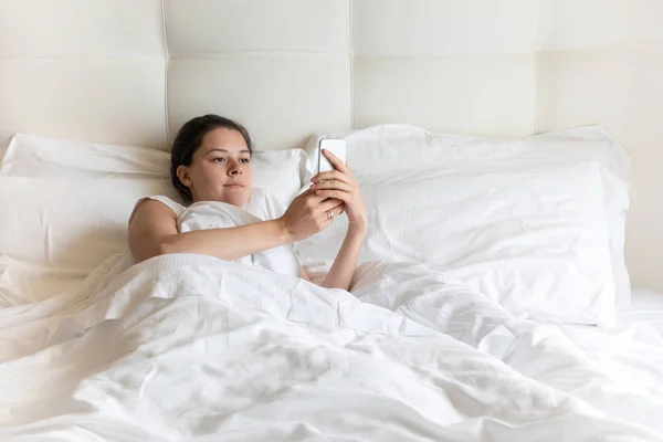 De mooie jonge meisje portret met slimme telefoon op bed in de ochtend — Stockfoto