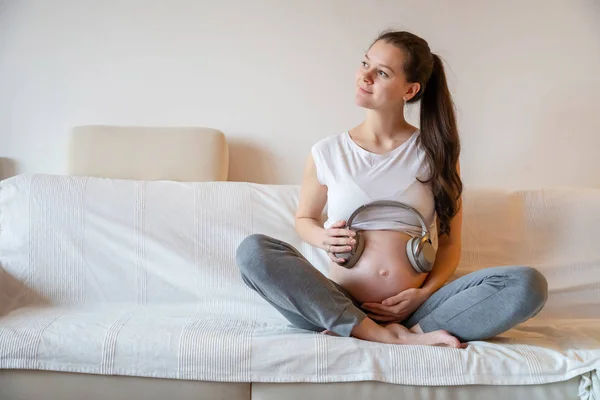 Těhotná žena sedí na bílé pohovce doma a drží sluchátka na břiše — Stock fotografie