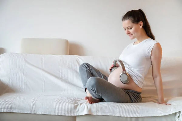 Schwangere sitzt zu Hause auf weißem Sofa und trägt Kopfhörer auf dem Bauch — Stockfoto