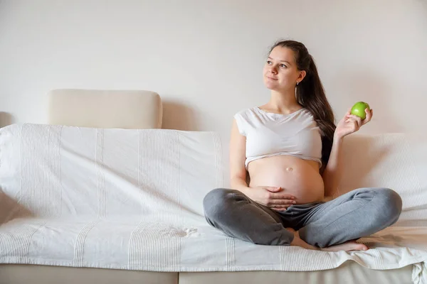 Junge schwangere Frau isst frischen grünen Apfel auf weißem Sofa — Stockfoto