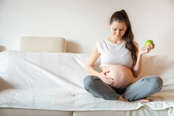 Junge schwangere Frau isst frischen grünen Apfel auf weißem Sofa — Stockfoto