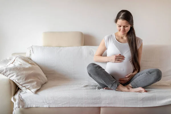 Ung gravid kvinna sitter på vit soffa och hålla händerna på magen — Stockfoto