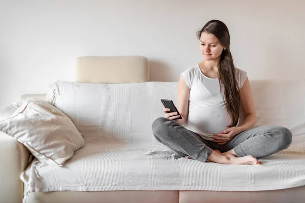 Jonge zwangere vrouw zit op een witte bank met smartphone en houdt de hand op de buik — Stockfoto