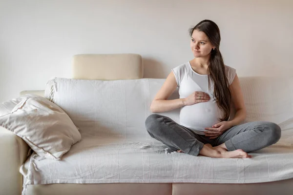 Junge schwangere Frau sitzt auf weißem Sofa und hält die Hände auf dem Bauch — Stockfoto