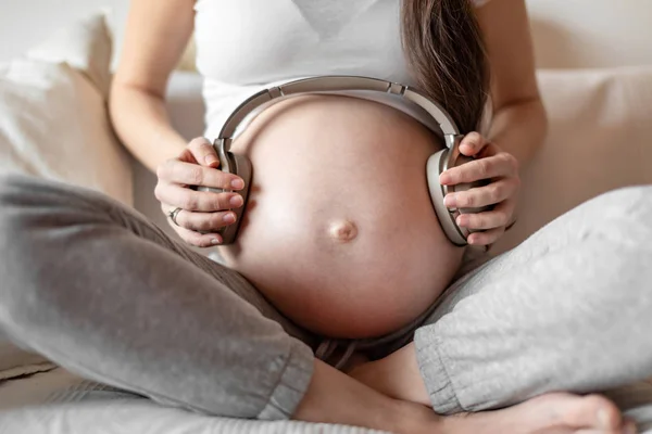 Mujer embarazada sentada en un sofá blanco y con auriculares en el vientre — Foto de Stock