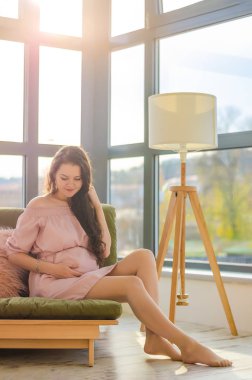 Genç hamile kadın güneşli bir sabahın tadını çıkarıyor. Evdeki büyük pencerenin yanında.