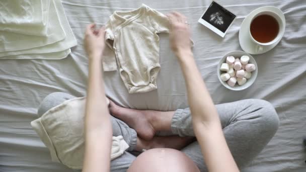 ベッドの中で妊娠中の女性のトップビュー自宅で赤ちゃんの服を準備 妊娠と出産の概念 — ストック動画