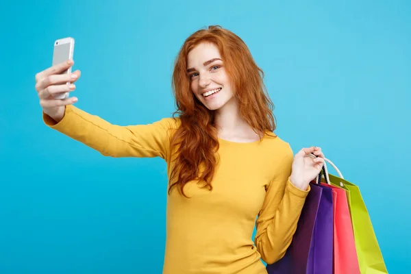Nákupní koncept - zblízka portrét mladé krásné redhair atraktivní dívka, usmívající se, při pohledu na fotoaparát s bílým nákupní tašku a selfie. Pastelové modré pozadí. Kopírovat prostor. — Stock fotografie