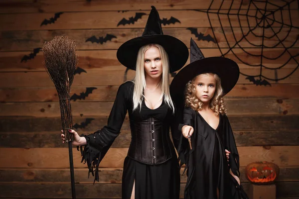 Хэллоуин Концепция - веселая мать и ее дочь в костюмах ведьм празднует Хэллоуин позируя с изогнутыми тыквы над летучих мышей и паутина на фоне деревянной студии . — стоковое фото