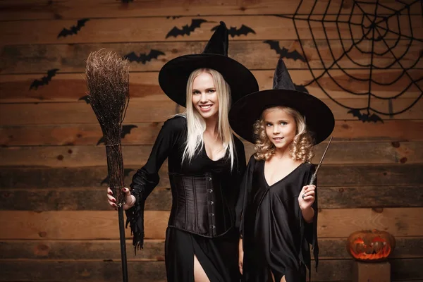 Halloween Concept - allegra madre e sua figlia in costumi da strega che celebrano Halloween in posa con zucche curve su pipistrelli e ragnatela su sfondo studio di legno . — Foto Stock