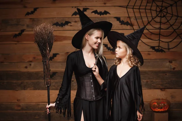 Concepto de Halloween - madre alegre y su hija en trajes de bruja celebrando Halloween posando con calabazas curvas sobre murciélagos y tela de araña sobre fondo de estudio de madera . —  Fotos de Stock