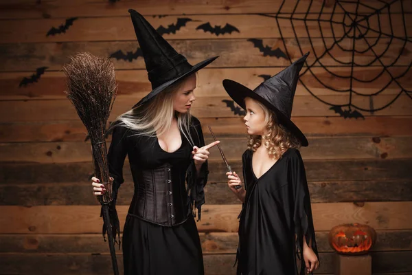 Concept Halloween - mère gaie et sa fille en costumes de sorcière célébrant Halloween posant avec des citrouilles courbes sur des chauves-souris et toile d'araignée sur fond de studio en bois . — Photo