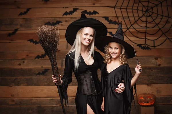 Concepto de Halloween - madre alegre y su hija en trajes de bruja celebrando Halloween posando con calabazas curvas sobre murciélagos y tela de araña sobre fondo de estudio de madera . —  Fotos de Stock