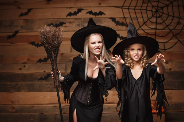 Concepto de Halloween - madre alegre y su hija en trajes de bruja celebrando Halloween posando con calabazas curvas sobre murciélagos y tela de araña sobre fondo de estudio de madera . — Foto de Stock