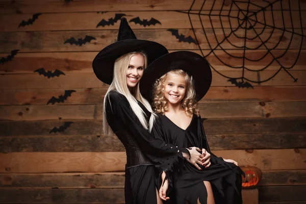 Halloween Concept - vrolijke moeder en haar dochter in heks kostuums vieren Halloween poseren met gebogen pompoenen over vleermuizen en spinnenweb op houten studio achtergrond. — Stockfoto