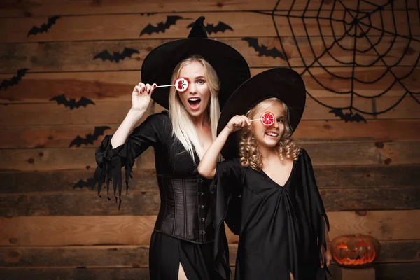 Halloween-konceptet - vackra kaukasiska mor och hennes dotter i häxa kostymer firar Halloween med Halloween godis och söta över fladdermöss och spindelnät på trä studio bakgrund. — Stockfoto