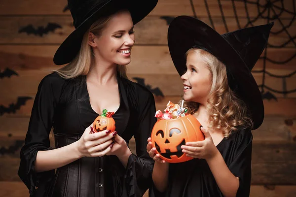 Halloween Concept - bella madre caucasica e sua figlia in costumi da strega che festeggiano Halloween con la condivisione di caramelle di Halloween e dolci su pipistrelli e ragnatela su sfondo in legno studio . — Foto Stock