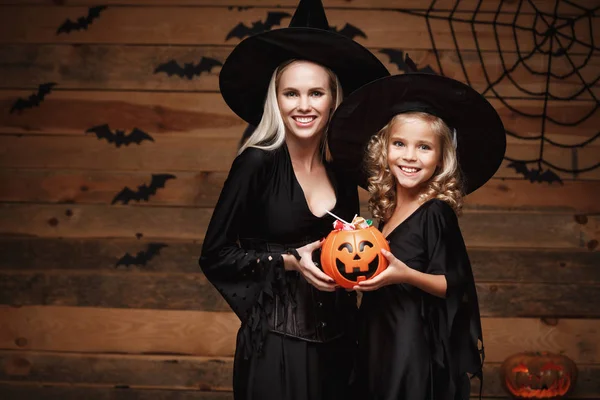 Halloween Concept - hermosa madre caucásica y su hija en trajes de bruja celebrando Halloween con el intercambio de dulces de Halloween y dulce sobre los murciélagos y la tela de araña en el fondo del estudio de madera . — Foto de Stock