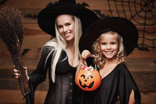 Halloween-konceptet - vackra kaukasiska mor och hennes dotter i häxa kostymer firar Halloween med delad Halloween godis och söta över fladdermöss och spindelnät på trä studio bakgrund. — Stockfoto