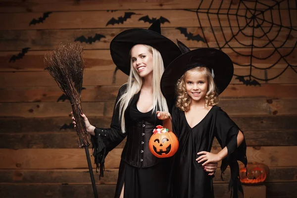 Halloween Concept - hermosa madre caucásica y su hija en trajes de bruja celebrando Halloween con el intercambio de dulces de Halloween y dulce sobre los murciélagos y la tela de araña en el fondo del estudio de madera . — Foto de Stock