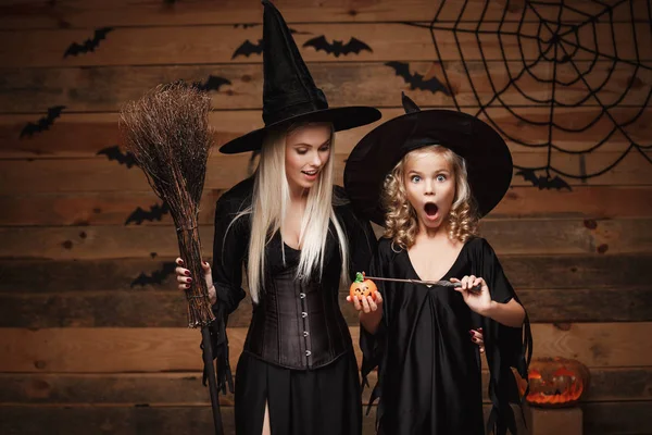 Halloween-Konzept - schöne kaukasische Mutter und ihre Tochter in Hexenkostümen genießen es, mit Zauberstab zu Halloween Kürbiskrüge über Fledermäuse und Spinnennetz auf hölzernem Studiohintergrund zu zaubern. — Stockfoto