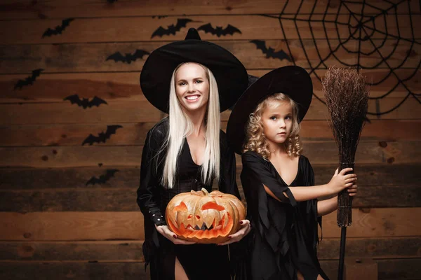 Concepto de Halloween - madre alegre y su hija en trajes de bruja celebrando Halloween posando con calabazas curvas sobre murciélagos y tela de araña sobre fondo de estudio de madera . — Foto de Stock