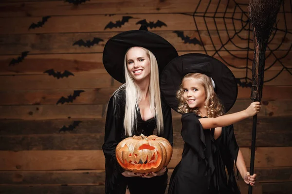 Cadılar Bayramı kavramı - neşeli anne ve kızı cadı kostümleri Cadılar Bayramı ile eğri kabaklar yarasalar ve örümcek ağı ahşap studio arka plan üzerinde poz kutluyor.