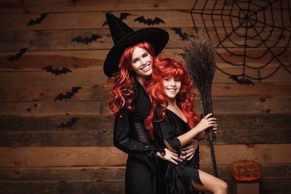 Concepto de Halloween - Hermosa madre caucásica y su hija con el pelo largo y rojo en trajes de bruja celebrando Halloween posando con más de murciélagos y tela de araña sobre fondo de estudio de madera . — Foto de Stock