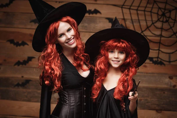 Koncept Halloween - krásné Kavkazský matka a její dcera s dlouhé červené vlasy v čarodějnice kostýmy a kouzelná hůlka slaví Halloween pózuje s netopýry a pavučina na dřevěné studio pozadí. — Stock fotografie