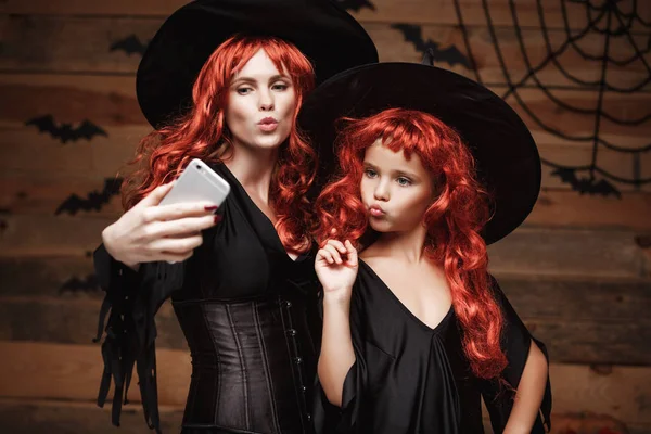 Halloween Concept - mooie Kaukasische moeder en haar dochter met lange rode haren in heks kostuums nemen een selfie met smartphone klaar voor het vieren van Halloween. — Stockfoto