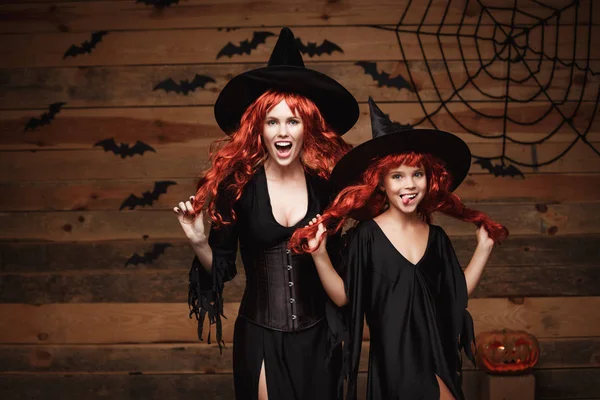 Концепция Хэллоуина - красивая кавказская мать и ее дочь с длинными рыжими волосами в костюмах ведьм празднуют Хэллоуин позируя с над летучими мышами и паутиной на фоне студии Wooden . — стоковое фото