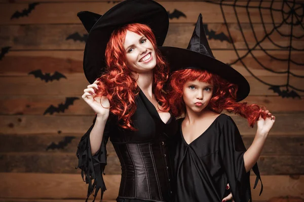 Halloween Concept - mooie Kaukasische moeder en haar dochter met lange rode haren in heks kostuums vieren Halloween poseren met over vleermuizen en spinnenweb op houten studio achtergrond. — Stockfoto