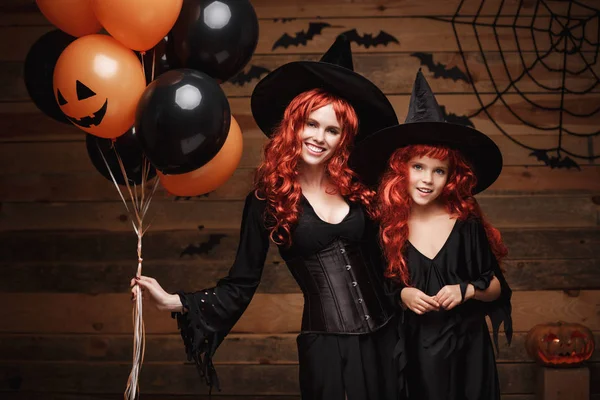 Halloween heks Concept - vrolijke moeder en haar dochter in heks kostuums vieren Halloween poseren met oranje en zwarte ballon over vleermuizen en spinnenweb op houten studio achtergrond. — Stockfoto