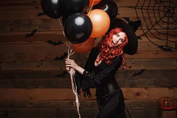Halloween heks Concept - mooie Kaukasische vrouw in heks kostuums vieren Halloween poseren met poseren met oranje en zwarte ballon over vleermuizen en spinnenweb op houten studio achtergrond. — Stockfoto
