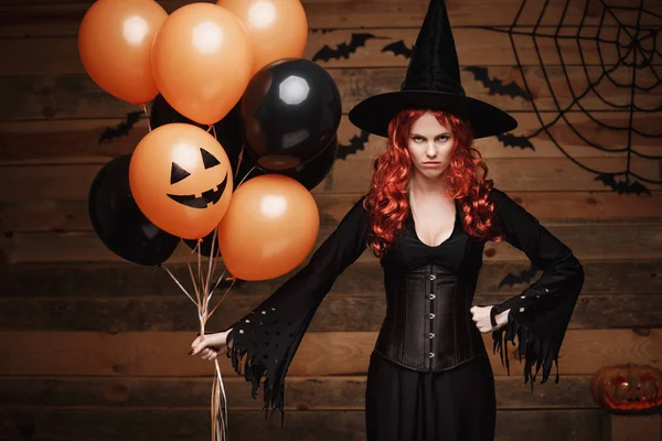 Halloween heks Concept - mooie Kaukasische vrouw in heks kostuums vieren Halloween poseren met poseren met oranje en zwarte ballon over vleermuizen en spinnenweb op houten studio achtergrond. — Stockfoto