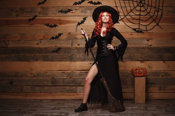 Koncepcja witch halloween - pełnometrażowych Happy Halloween czerwone włosy czarownica trzyma pozowanie z magicznej miotły nad stare drewniane studio tło. — Zdjęcie stockowe