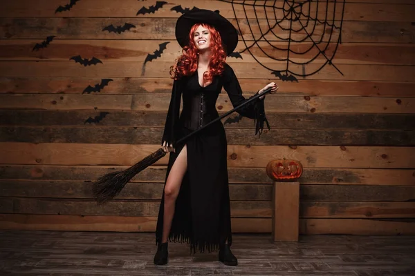 Halloween heks concept - Full-length Happy Halloween rode haren Witch holding poseren met magische bezemsteel op oude houten studio achtergrond. — Stockfoto