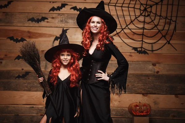 Halloween concept - schöne kaukasische Mutter und ihre Tochter mit langen roten Haaren in Hexenkostümen feiern halloween posieren mit Fledermäusen und Spinnennetz auf hölzernem Studiohintergrund. — Stockfoto