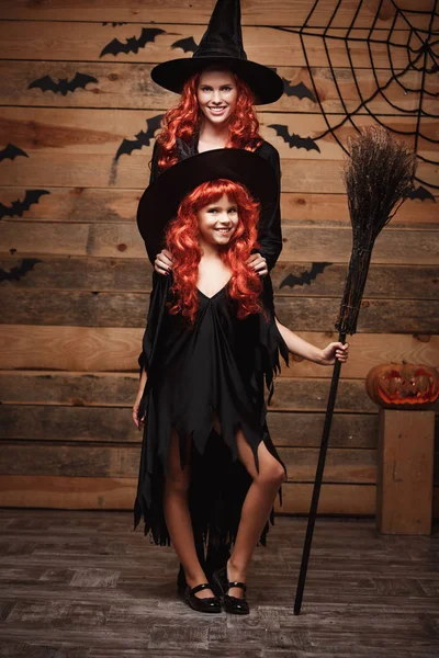 Halloween Concept - mooie Kaukasische moeder en haar dochter met lange rode haren in heks kostuums vieren Halloween poseren met over vleermuizen en spinnenweb op houten studio achtergrond. — Stockfoto
