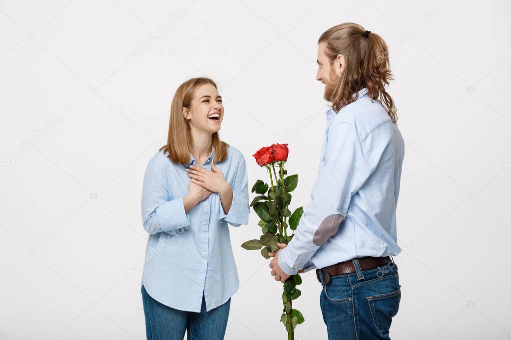 Portrait of handsome elegant guy is surprising his beautiful girlfriend with red roses and smiling over white isolated background.