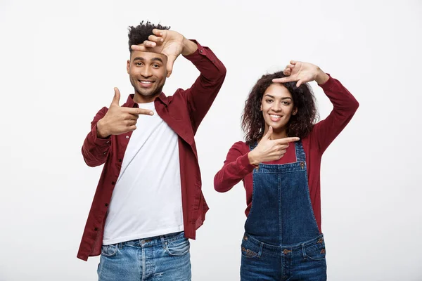 Feliz joven pareja afroamericana mirando a través de un marco de dedo y sonriendo mientras está de pie aislado en blanco . —  Fotos de Stock